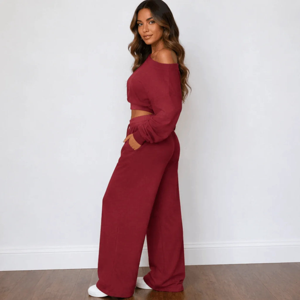 A woman modeling a matching red two-piece loungewear set consisting of an off-the-shoulder long-sleeve crop top and wide-leg pants, standing against a white wall.