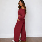 A woman modeling a matching red two-piece loungewear set consisting of an off-the-shoulder long-sleeve crop top and wide-leg pants, standing against a white wall.