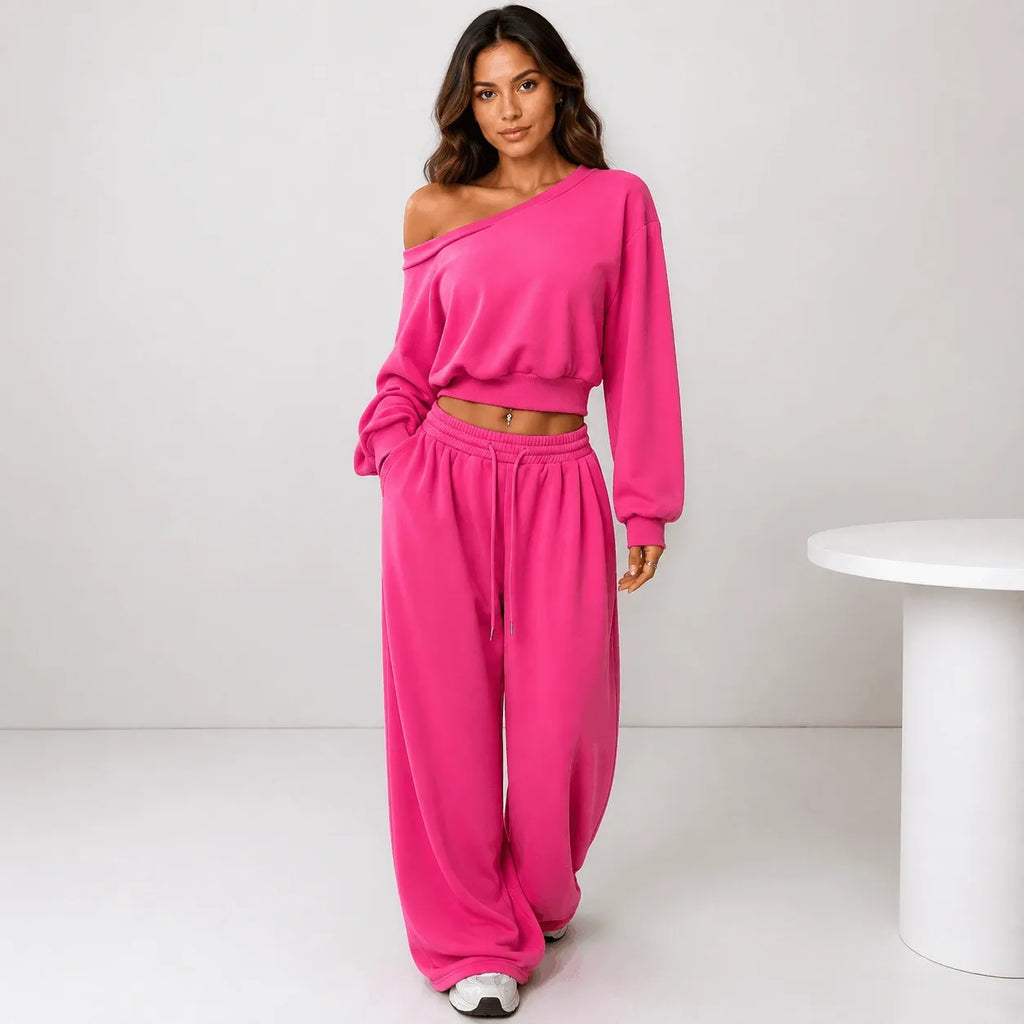 A woman modeling a bright pink two-piece lounge set consisting of an asymmetrical long-sleeve crop top and matching wide-leg sweatpants, standing in a minimalist studio setting.
