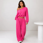 A woman modeling a bright pink two-piece lounge set consisting of an asymmetrical long-sleeve crop top and matching wide-leg sweatpants, standing in a minimalist studio setting.