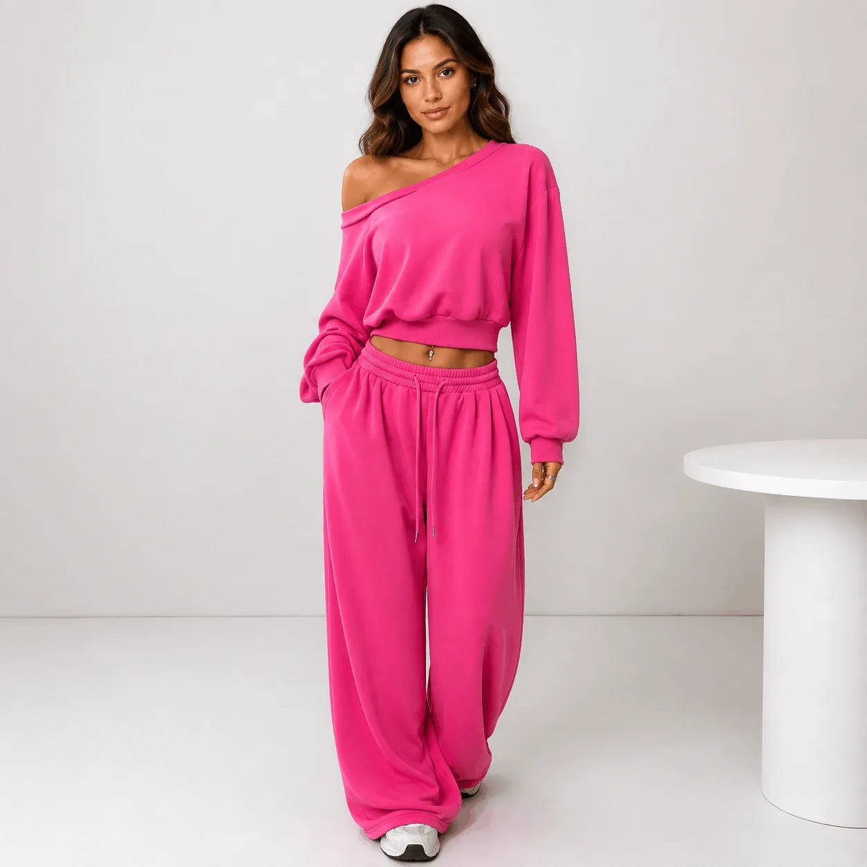 A woman modeling a bright pink two-piece lounge set consisting of an asymmetrical long-sleeve crop top and matching wide-leg sweatpants, standing in a minimalist studio setting.