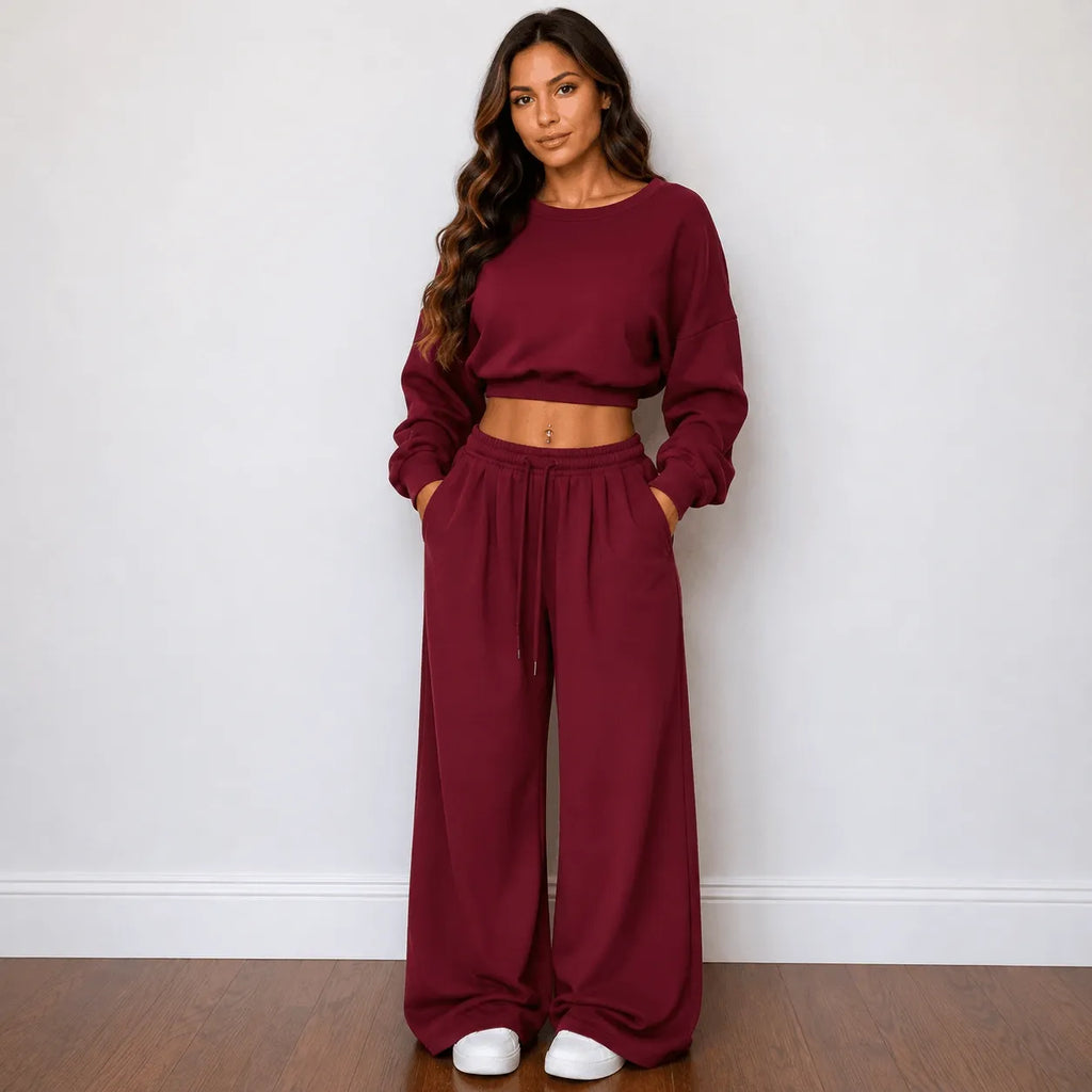 A woman modeling a matching burgundy two-piece lounge set consisting of a long-sleeve cropped sweatshirt and wide-leg drawstring pants, standing against a white wall on a wooden floor.