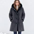 A woman wearing a dark gray hooded shearling-style coat with toggle closures, standing against a bright white snowy background.