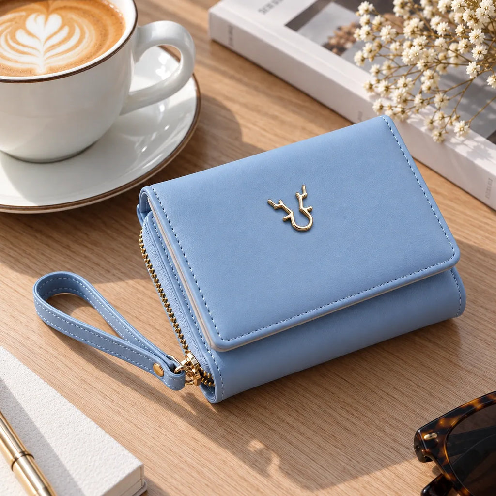 A light blue faux-leather wallet with a wrist strap and gold deer logo, styled on a wooden table next to a coffee cup, book, and sunglasses.