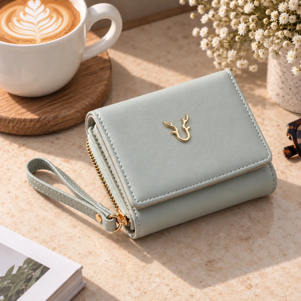 A light sage green wallet with a wrist strap and gold deer emblem, styled on a beige tabletop next to a cup of latte and dried flowers.