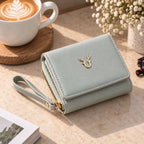 A light sage green wallet with a wrist strap and gold deer emblem, styled on a beige tabletop next to a cup of latte and dried flowers.