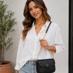 Women's black faux leather shoulder bag with gold chain strap and magnetic flap closure styled with a white button-down shirt and light wash jeans.