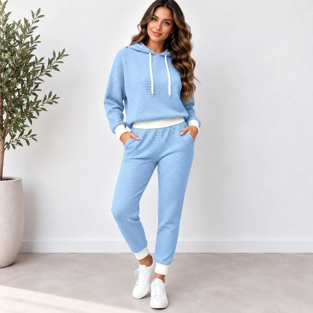 A woman modeling a light blue waffle-knit two-piece loungewear set consisting of a cropped hoodie and matching jogger pants, styled with white sneakers in a bright indoor setting.