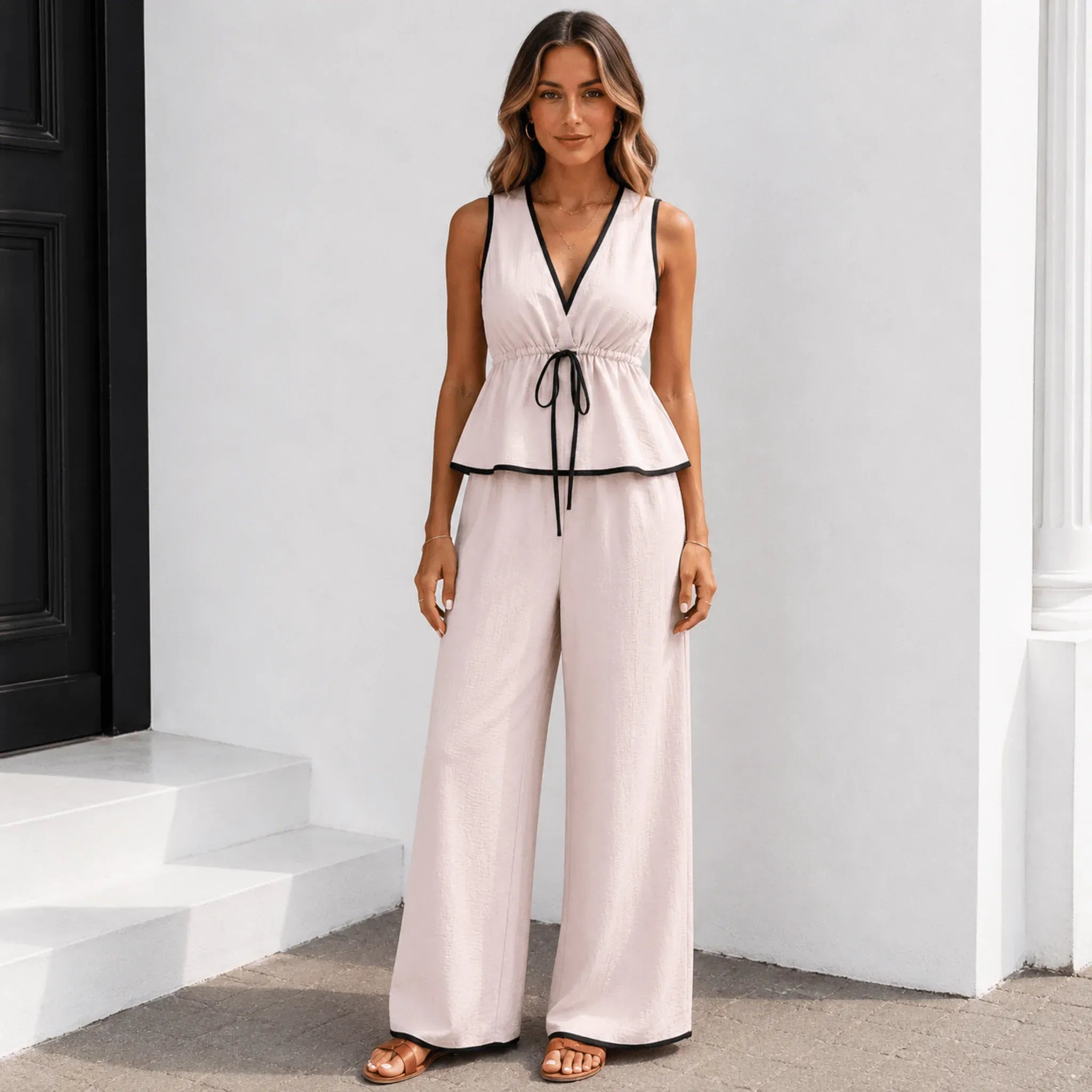 Women's sleeveless two-piece set in blush pink with black trim featuring a peplum top and wide-leg pants.