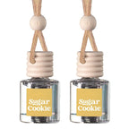 Two small clear glass reed diffusers with pale wood caps, beige hanging cords, and yellow labels reading 'Sugar Cookie' on a white background.