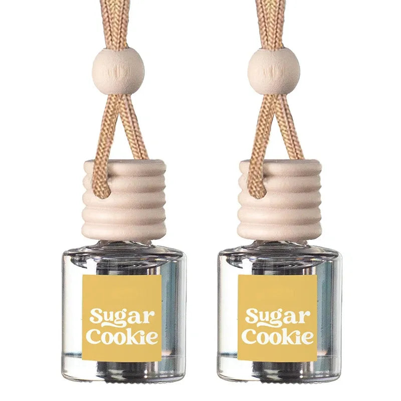 Two small clear glass reed diffusers with pale wood caps, beige hanging cords, and yellow labels reading 'Sugar Cookie' on a white background.
