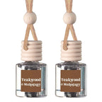 Two identical reed diffuser bottles with clear glass bodies, beige wooden stoppers, and natural rope hangers. Each bottle has a brown label reading 'Teakwood & Mahogany' in white text, set against a plain white background.