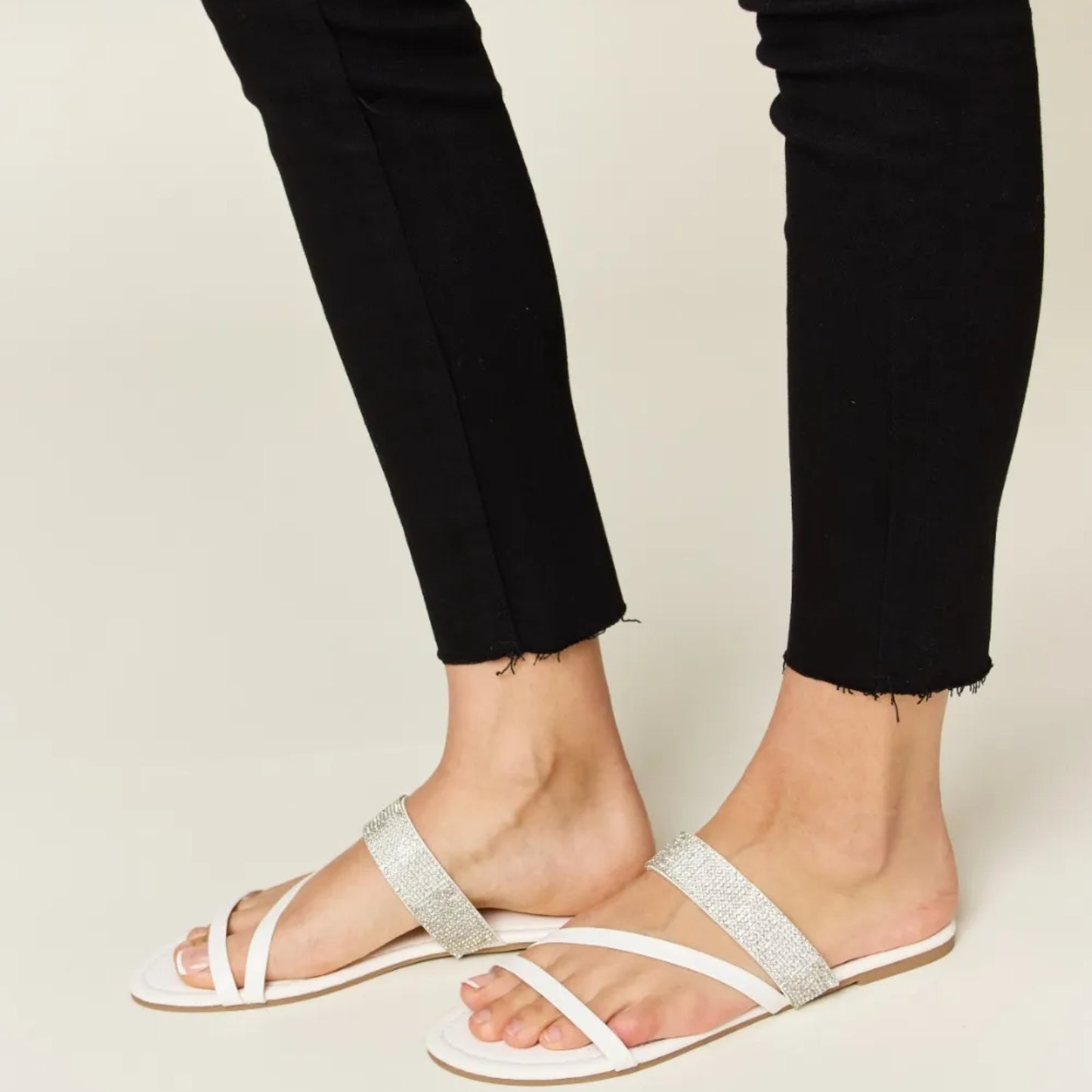 White sandals with glittery straps worn with black pants on a neutral background