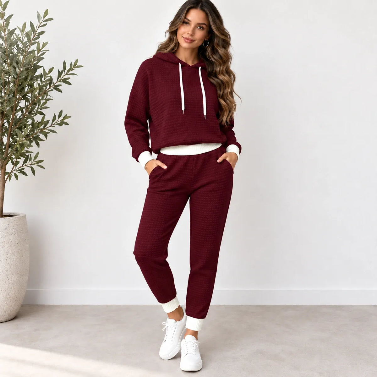 A woman modeling a matching two-piece lounge set consisting of a cropped waffle-knit hoodie and jogger pants in burgundy, with white drawstrings, white ribbed cuffs and hem, and white sneakers, standing in a bright room with a potted plant.