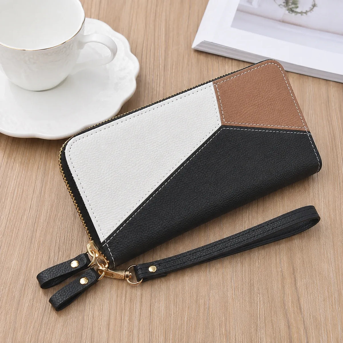 A color-blocked zip-around wallet with a wrist strap, placed on a light wood tabletop next to a white teacup and saucer.