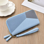 A light blue and gray geometric zip-around wallet with a wrist strap, displayed on a wooden tabletop next to a white teacup and saucer.