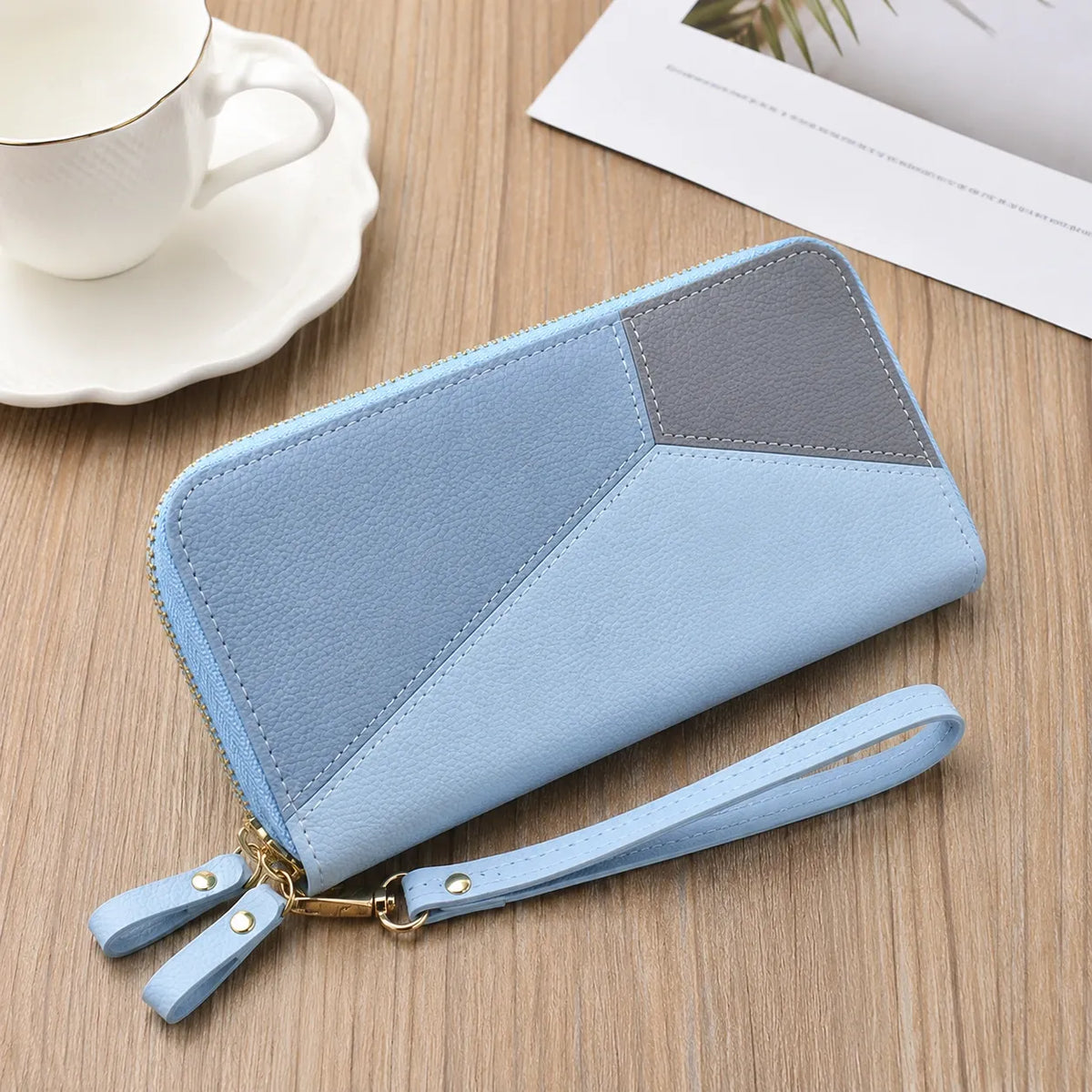 A light blue and gray geometric zip-around wallet with a wrist strap, displayed on a wooden tabletop next to a white teacup and saucer.