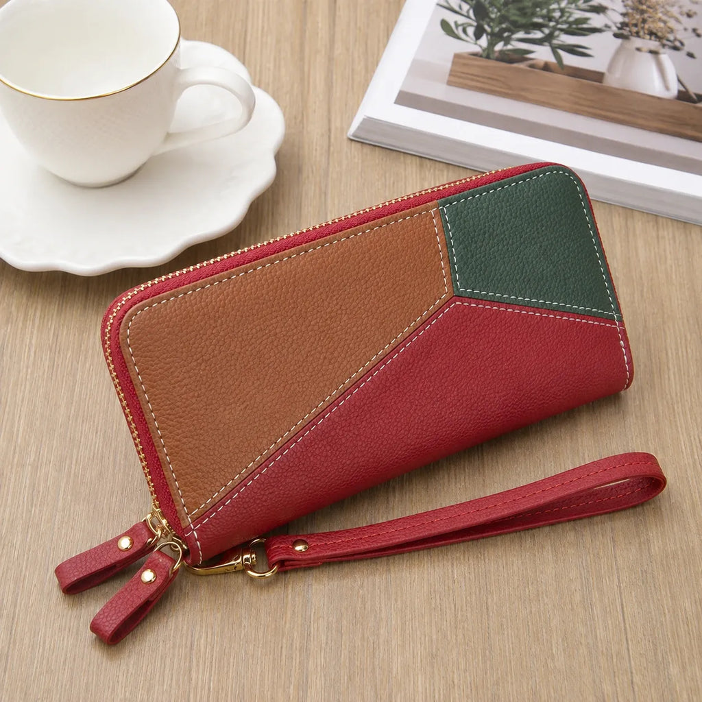A geometric patchwork leather zip-around wallet with a wrist strap, styled on a light wood tabletop next to a white teacup and saucer.