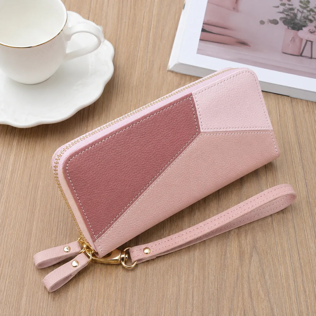 A pink geometric zip-around wallet with a wrist strap and gold-tone hardware, photographed on a wooden tabletop with a white cup and saucer and a framed print in the background.
