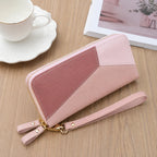A pink geometric zip-around wallet with a wrist strap and gold-tone hardware, photographed on a wooden tabletop with a white cup and saucer and a framed print in the background.