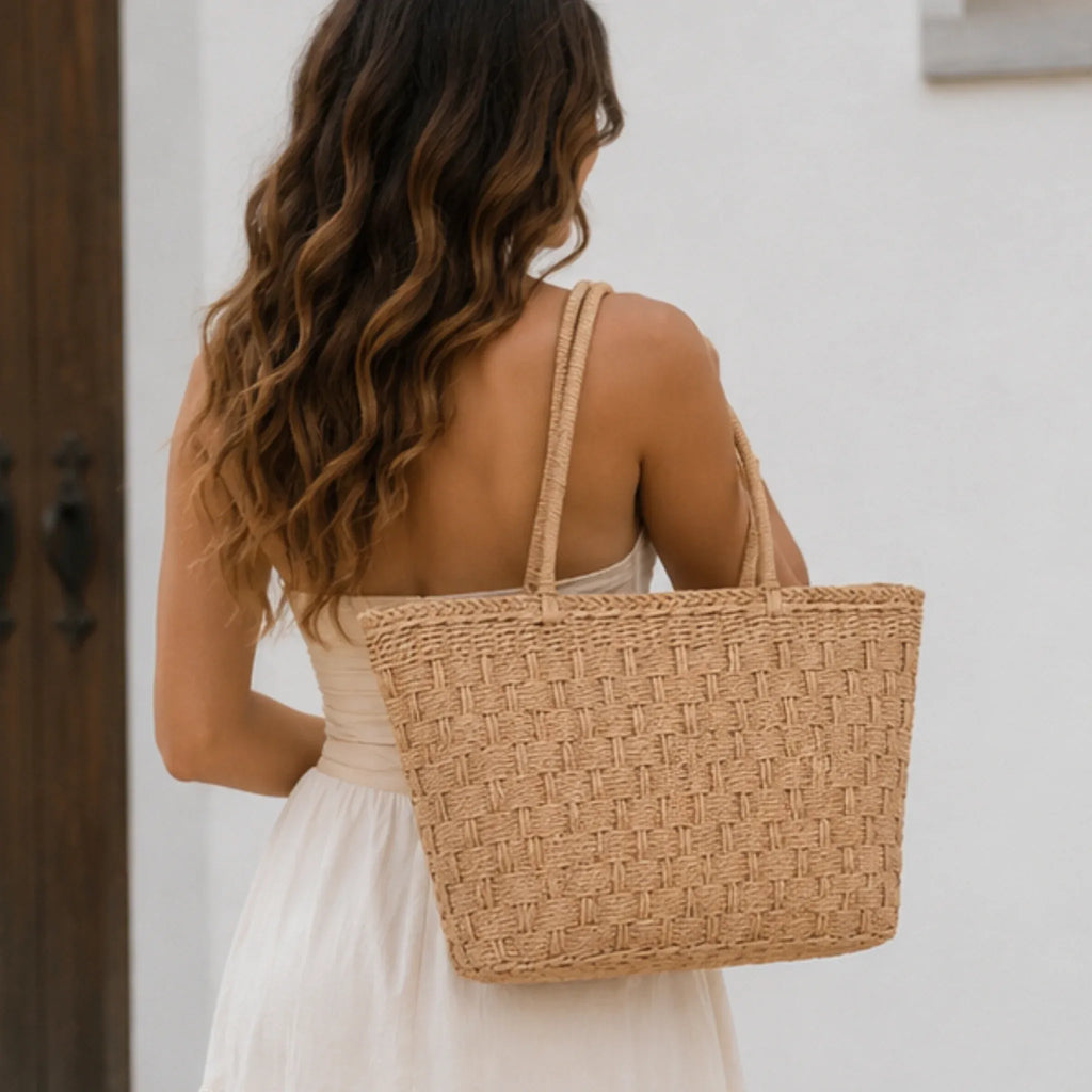 Back view of a woven straw tote bag with double handles worn over the shoulder with a white summer dress.